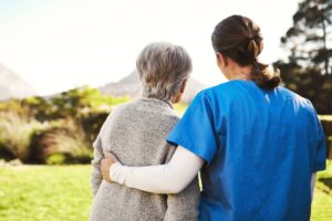 A Home Care Aide with her arm around a senior woman as they both are standing outside looking away from the camera