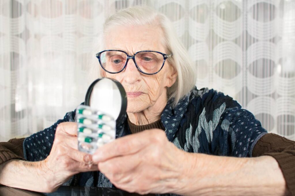 An elderly woman with big glasses using a magnifying glass to read a pill label