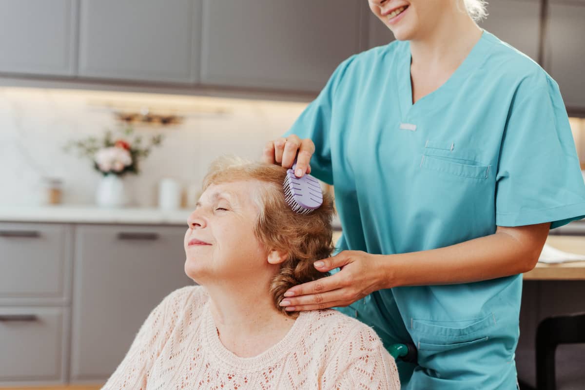 A senior woman is having her hair brushed by a caregiver